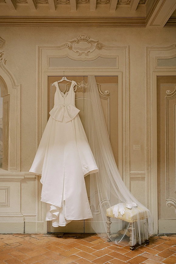 Wedding dress hanging with bridal dress hanging detail, veil draped and gloves on an upholstered chair in an ornate bridal suite interior