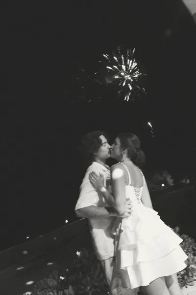 Wedding kiss portrait during a wedding fireworks moment, newlyweds embrace under a night sky with garden foliage softly aglow.