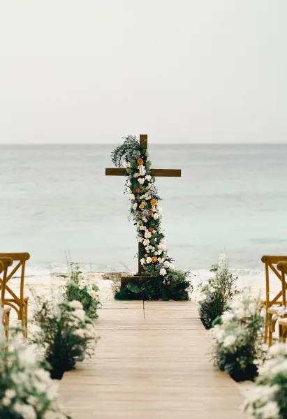 Wedding cross altar with white and peach florals anchors beach ceremony decor on a wooden boardwalk, framed by ocean waves and open sky.
