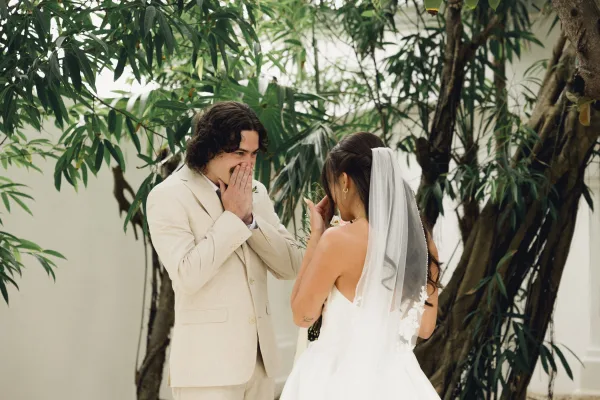 Wedding first look, a groom reaction photo as the bride wipes tears beneath a long veil, framed by garden trees against a white wall.