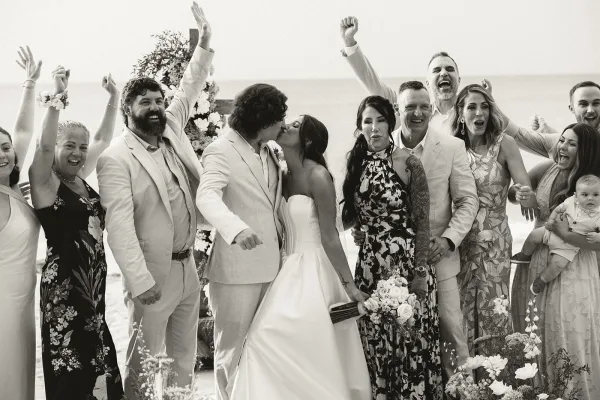 Group wedding portrait with wedding party cheering beneath a floral arch on a sandy beach, the ocean and bright sky framing the scene.