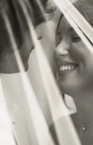 Couple portrait in black and white, faces nearly touching beneath a sheer veil as the bride smiles at the groom.