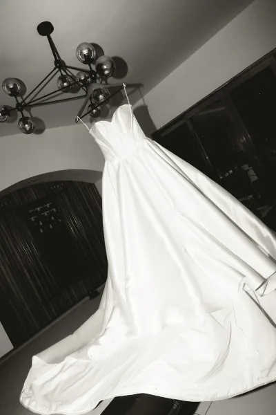 Wedding dress, a ballgown wedding dress, hangs on a hanger beneath a chandelier by arched windows, light pooling across the floor.