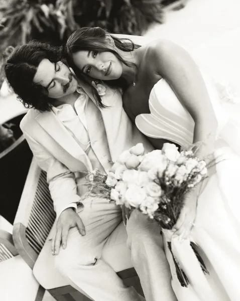 Couple portrait with bride leaning on groom as they sit on a patio bench, her rose bouquet bright against lush garden foliage.