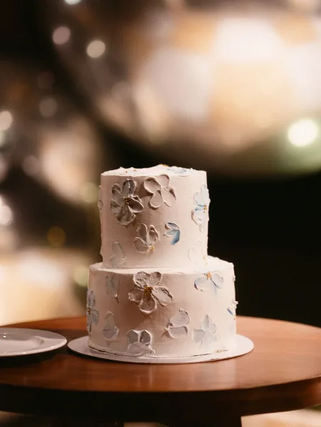 Wedding cake, a two tier wedding cake with textured white buttercream and delicate buttercream flowers on a wood table amid blurred lights.