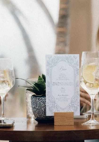 Bar menu sign for wedding bar signage, beside wine glasses, glowing with window light and softened by blurred foliage.