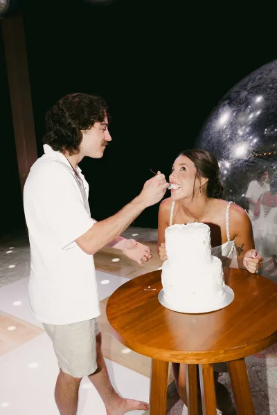 Cake cutting as the groom feeds the bride cake, both laughing on the dance floor under a shimmering disco ball and stark spotlights.