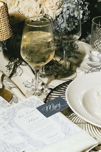 Wedding place setting with a calligraphy place card on a charger plate, framed by a floral arrangement over patterned table linen.