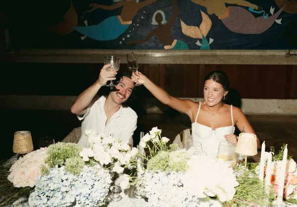 Reception toast with the bride and groom toasting at their sweetheart table, taper candles glowing warmly against a mural backdrop.