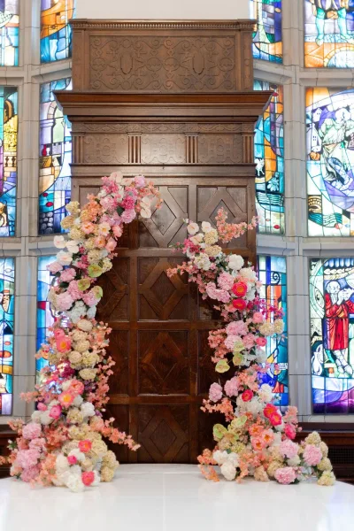 Wedding ceremony backdrop with a floral ceremony arch of peonies, hydrangeas, and orchids set before stained glass in a church interior