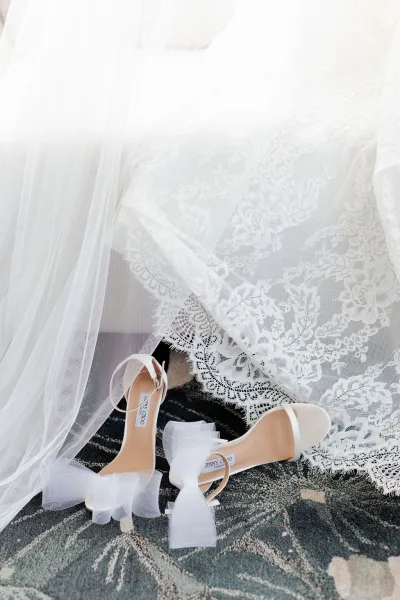 Bridal shoes with tulle bows in a wedding shoes flat lay beside lace dress hem and veil on patterned carpet background