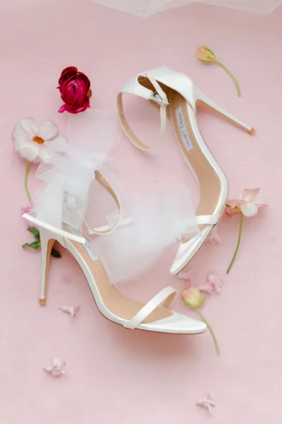 Wedding shoes with bridal heels in white strappy ankle-strap sandals, tulle bows, and flower petals on a pink sheer fabric backdrop