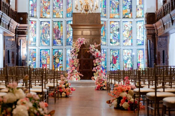Ceremony altar decor with a wedding floral arch and aisle florals beneath a chandelier, framed by stained glass and wood-paneled walls