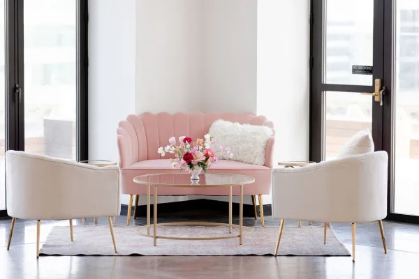 Wedding lounge seating with a pink velvet sofa, white chairs, and glass coffee table with floral centerpiece in a bright windowed room