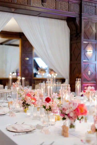 Reception tablescape with wedding table centerpieces, low pink and coral florals, floating candles in cylinder vases, and chiavari chairs in a wood-paneled ballroom