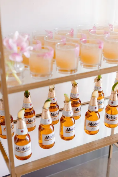 Wedding welcome drinks arranged on a glass shelf display with cocktails, beer bottles, lime wedges, and a pink flower garnish on white wall backdrop