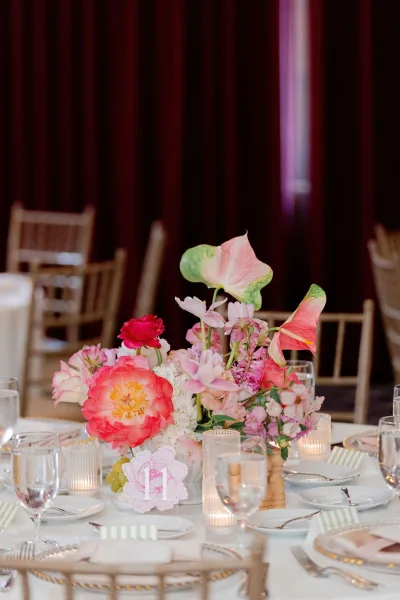 Reception tablescape with a wedding table centerpiece of calla lilies and peonies, gold rim charger plates, striped napkins, and candles in a dark-draped room