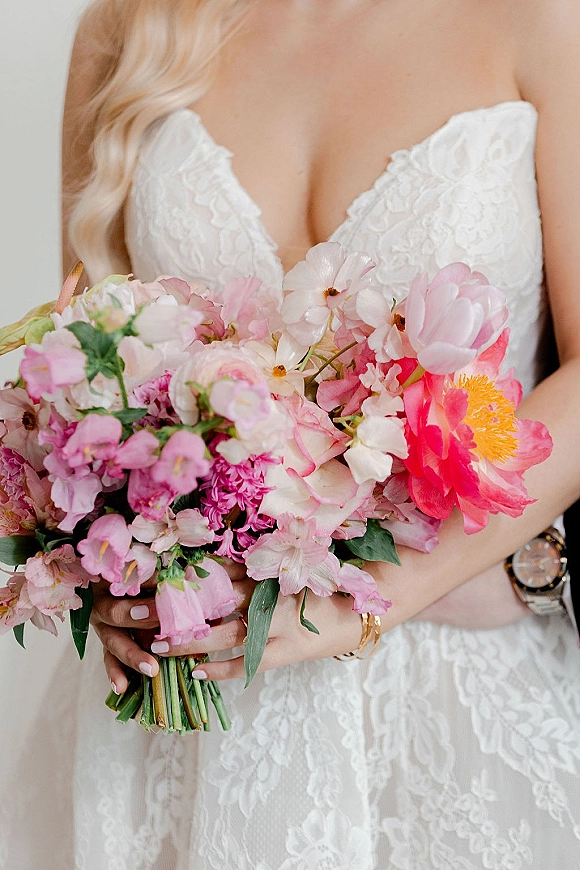 Bridal bouquet of pink peonies and greenery held against a strapless lace wedding dress, with bracelet and wristwatch details
