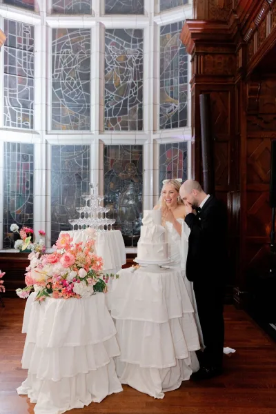 Cake cutting by the bride and groom at a white wedding cake beside a champagne tower, framed by stained glass windows and ruffled linens
