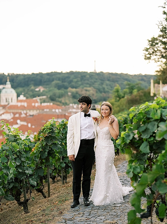 Couple portrait in a vineyard wedding portrait, bride in a strapless lace gown and groom in a white tuxedo walking on a cobblestone path
