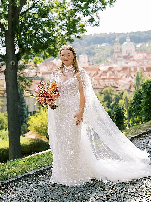 Bridal portrait of a bride holding bouquet in a lace wedding dress with a sheer cape veil on a cobblestone path overlooking city domes