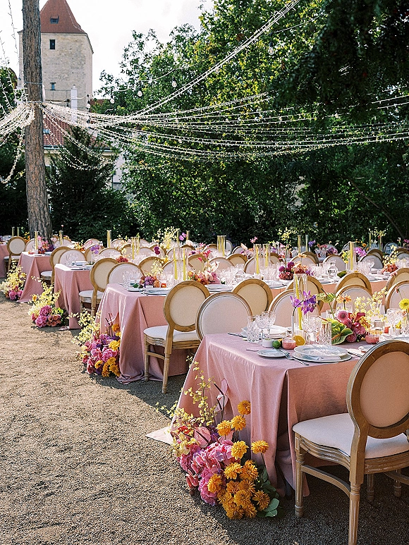Reception tablescape with pink linens, colorful floral centerpieces, taper candles and citrus on long tables under string lights in a garden courtyard