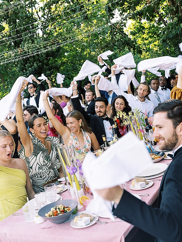 Wedding reception toast with guests cheering and waving white napkins over a long pink banquet table under string lights in a garden setting
