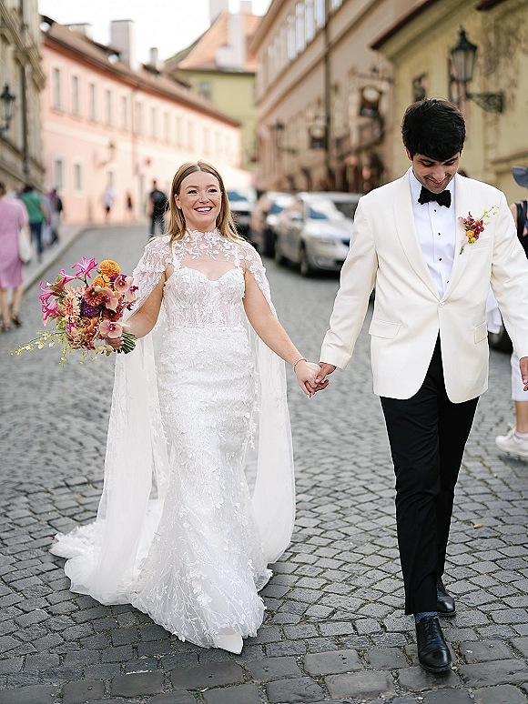Couple portrait of bride and groom walking hand in hand on a cobblestone street, bride holding a colorful bouquet, groom in white dinner jacket.