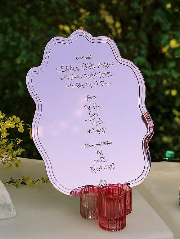 Bar menu sign with wedding bar menu in calligraphy on a pink-tinted scalloped acrylic plaque, flanked by pink ribbed glass votives in a garden setting