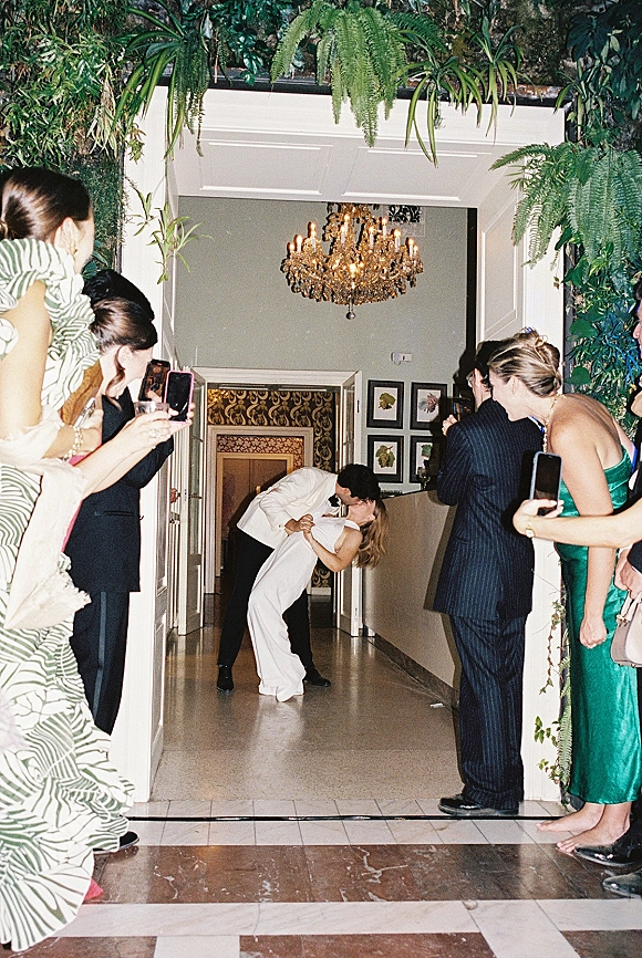 Wedding kiss moment as newlyweds dip kiss in a doorway under a crystal chandelier, guests lining the hallway cheering and filming on phones