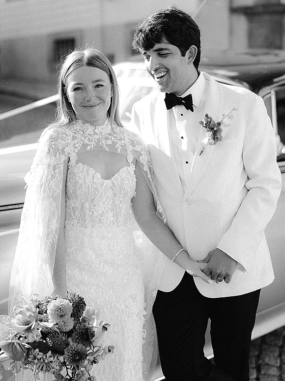 Couple portrait in black and white, bride in lace gown and cape holding a bouquet, groom in white tuxedo by a vintage car on the street