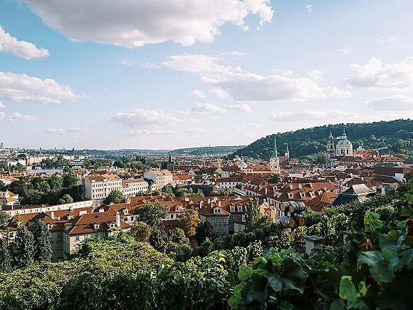 City skyline view over red tile rooftops and church towers from a hillside vineyard with wooden barrels under blue sky clouds