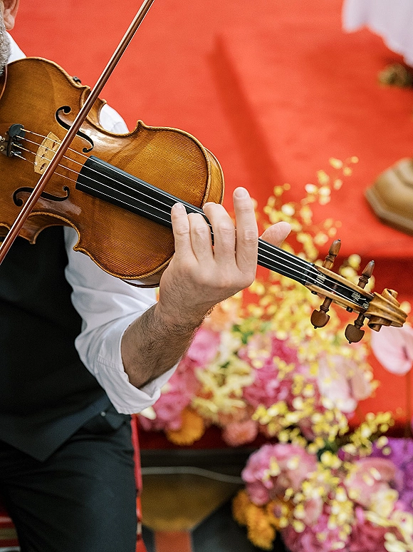 Wedding violinist playing ceremony live music with violin and bow, wearing a black vest on a red aisle runner with petals and florals