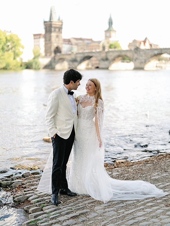 Couple portrait of bride and groom by river holding hands, her lace cape sleeves and long train flowing near a stone bridge backdrop