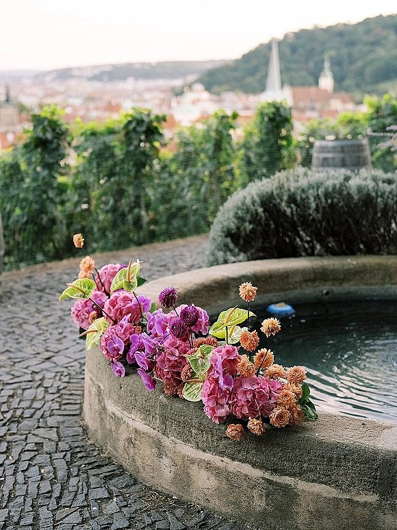 Wedding floral installation draped over a stone fountain with pink flower accents, purple and orange blooms and greenery on a cobblestone garden terrace
