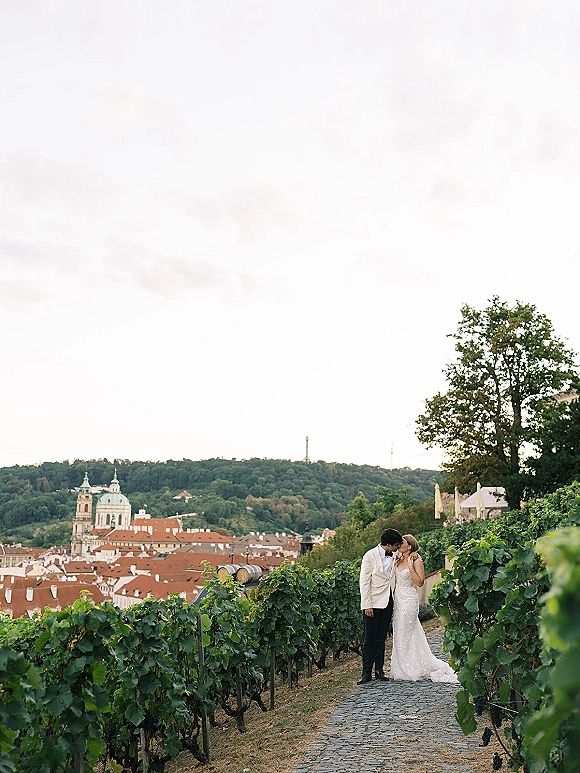 Wedding couple portrait of bride and groom kissing in vineyard wedding photos, her lace dress train flowing beside a cobblestone path with hillside town skyline