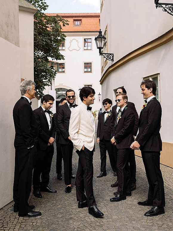 Groomsmen portrait of the groom and groomsmen in tuxedos, one in a white dinner jacket, walking on a cobblestone street past white stucco buildings