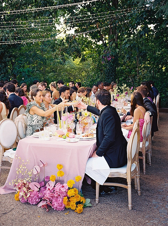 Reception dinner setup with a long banquet table dressed in blush pink tablecloth, florals and taper candles under string lights in a garden