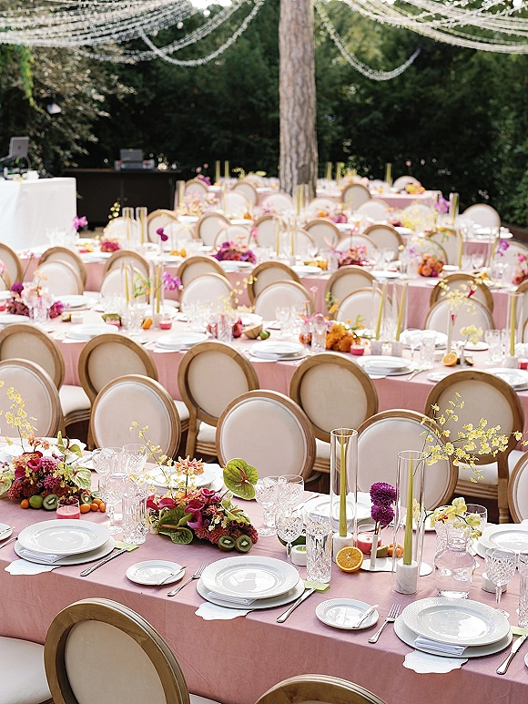 Reception tablescape with pink wedding table linens, floral centerpieces, citrus, taper candles and crystal glassware under garden string lights