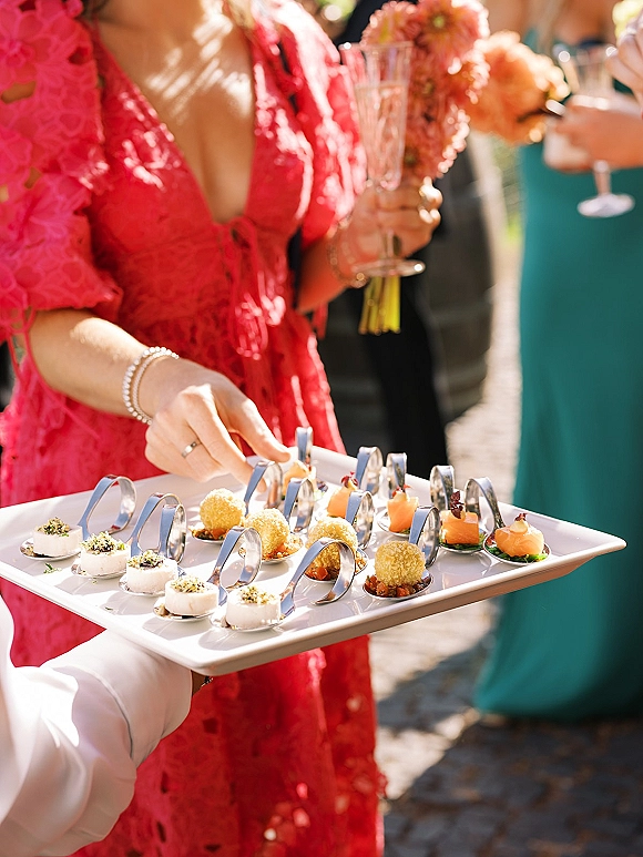 Wedding cocktail hour with passed hors d'oeuvres on an appetizer tray, guests holding champagne flutes and bouquets in an outdoor setting
