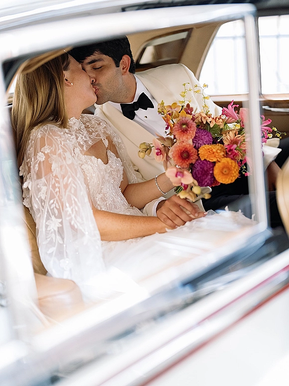 Wedding kiss between newlyweds in car, bride holding a colorful bouquet as groom in white tuxedo and bow tie leans in, veil draped