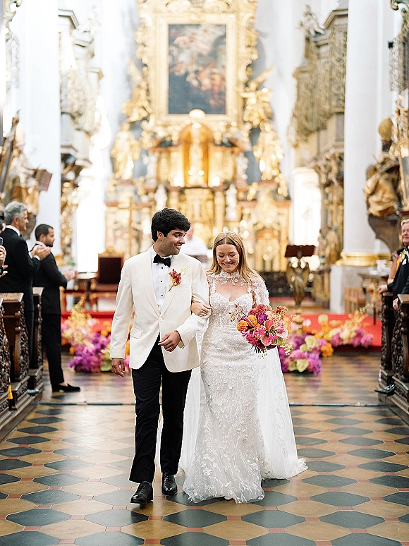 Wedding recessional as bride and groom walk the aisle, bride holding a pink-orange bouquet, past guests toward an ornate gold altar