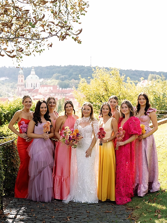 Bridesmaid group photo with bride with bridesmaids, all holding bouquets in colorful dresses on a cobblestone path in a garden hillside setting