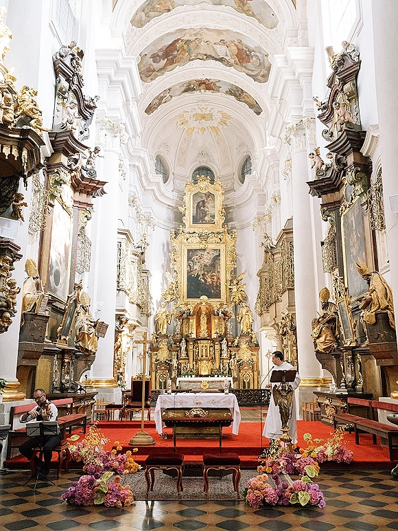 Church ceremony altar with a gold gilded altar backdrop, white cloth, candles and flowers, framed by baroque columns and fresco ceiling