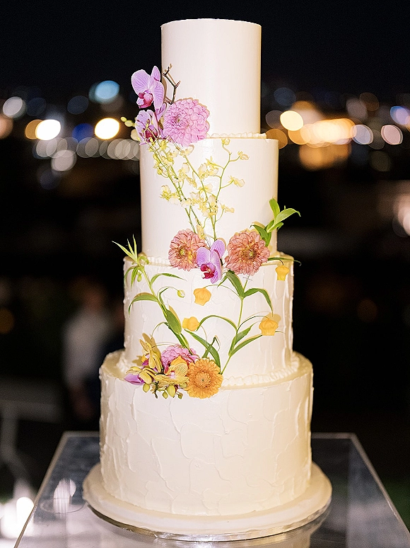 Wedding cake with tiered white buttercream frosting, orchids and dahlias with greenery on a cake stand against nighttime bokeh lights