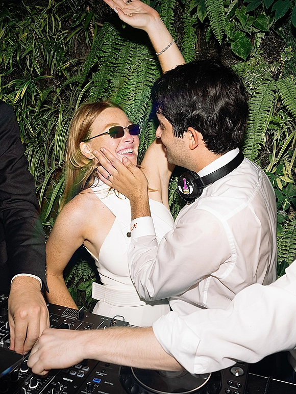 Wedding reception moment at a wedding dj booth with bride in sunglasses and groom in headphones by a lush greenery wall backdrop
