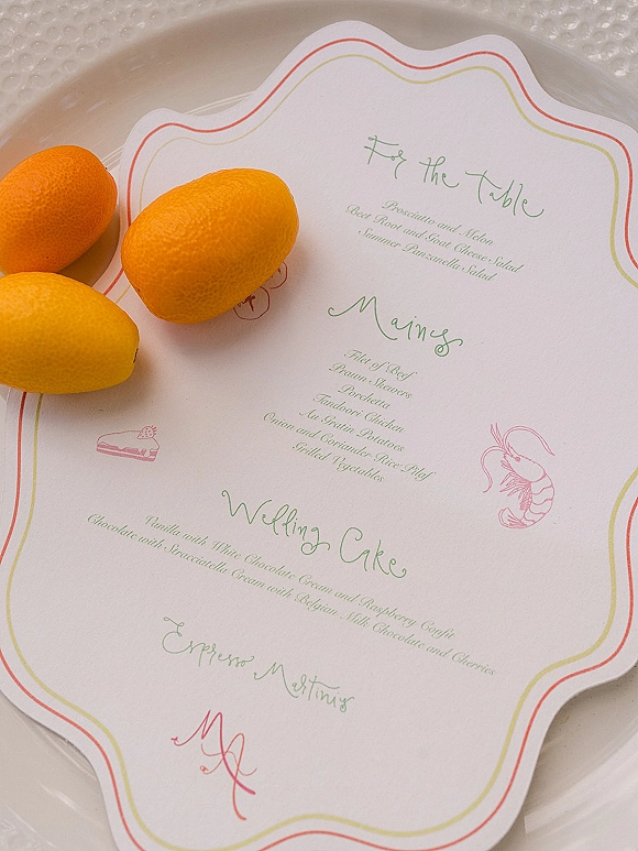Wedding menu card with green calligraphy and red-yellow border, illustrated shrimp and cake, resting on a white textured plate flat lay