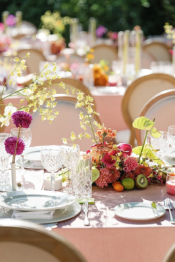 Reception tablescape with pink wedding table decor, bright floral and fruit centerpiece, candles, crystal glassware, and bud vases in garden light