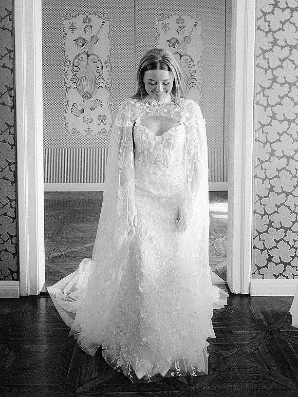 Bridal portrait in black and white of a bride looking down in a long sleeve lace gown with sheer cape, framed by ornate wallpaper doorway
