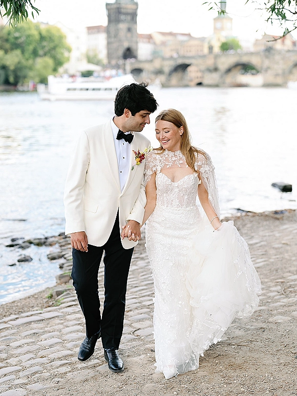 Couple portrait of bride and groom holding hands, her lace wedding dress with sheer cape, walking by a river near a stone bridge